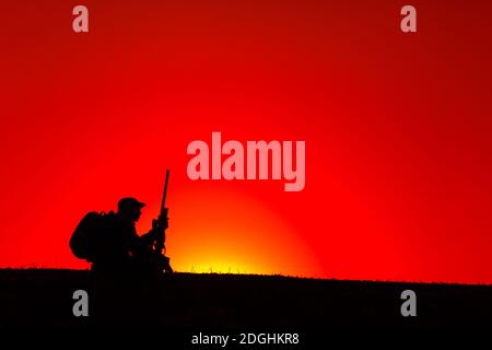 Silhouette de sniper militaire, de soldat des forces spéciales, de mercenaire professionnel ou de chasseur en bonnie, portant un sac à dos et assis au sommet de la colline avec un fusil de sniper sur fond de coucher de soleil ou de ciel de l'aube Banque D'Images