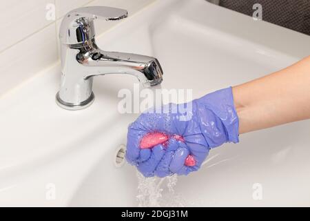 main féminine dans un gant bleu lave un chiffon sous le robinet d'eau dans la salle de bains. Nettoyage de la maison Banque D'Images