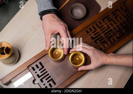 Vue de dessus d'un plateau en bambou avec un plateau. Les mains des hommes et des femmes se ferment en tenant des tasses en verre avec du thé ambré lors d'une cérémonie intime de thé Banque D'Images