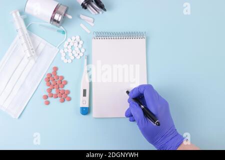 Médecine, pharmacie, concept de santé. Main du médecin dans le gant médical bleu écrire une prescription. Bureau bleu avec pilules, thermometr, masque médical, seringue Banque D'Images