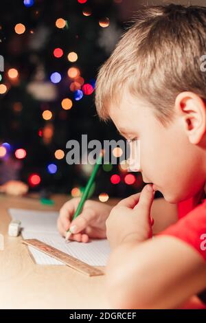 7 yeas vieux garçon assis à un bureau et faisant des devoirs. L'enfant écrit des notes dans le carnet, les devoirs difficiles, l'enfant se concentrant sur des exemples. Arbre de Noël décoré sur fond Banque D'Images