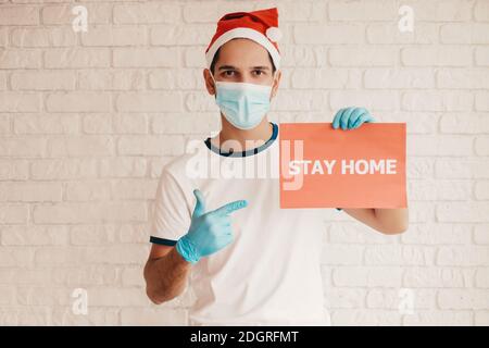 Joyeux homme médecin dans le chapeau de Santa, masque médical de visage, gants de protection pointe doigt sur la bannière rester à la maison texte. Jeune infirmière masculine dans le masque de visage montrer rester à Banque D'Images