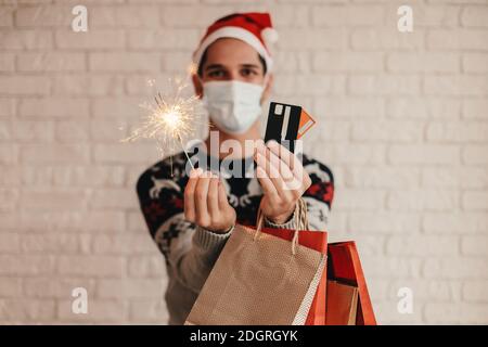Jeune homme festif en chandail de Noël, masque de protection célébrez la saison des soldes d'hiver. Happy man shopper en chapeau de père Noël, masque de maintien de la lumière de sparkler, c Banque D'Images