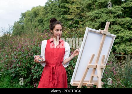 Fille souriante, belle femme en robe rouge, dessine l'image, en été sur la nature, arrière-plan arbres buissons fleurs. Peinture et pinceau pour chevalet Banque D'Images