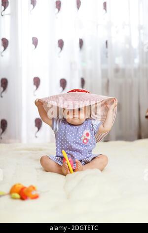 Un peu drôle caucasien petite fille un enfant s'assoit à la maison sur le sol sur un tapis clair. Sur sa tête est un grand chapeau d'été qui Banque D'Images