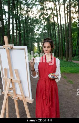 Fille peintre dans la forêt. Une femme en robe rouge, dessine une image, en été sur la nature dans le parc, arbres de fond. Peinture et pinceau pour chevalet Banque D'Images