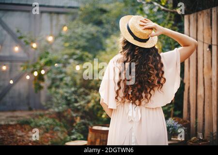 Une fille dans une longue robe rose et un chapeau de paille se tient avec son dos sur le fond de l'éclairage de rue sur des planches de bois. Banque D'Images