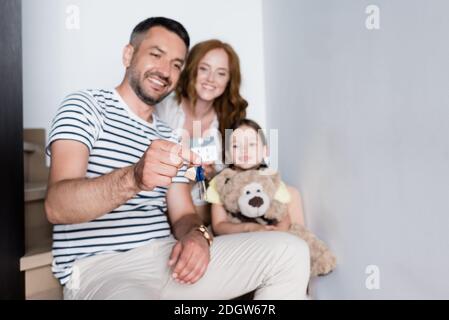 Fille souriante et femme regardant les clés dans les mains de homme assis dans les escaliers à la maison sur un arrière-plan flou Banque D'Images