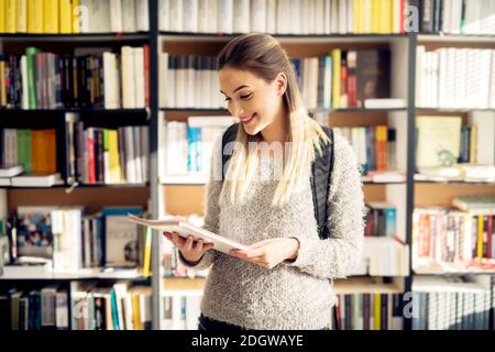 Portrait d'une jeune fille étudiante magnifique tenant et regardant son carnet tout en se tenant devant l'étagère de livres de bibliothèque. Banque D'Images