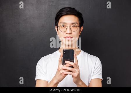 Technologie, messagerie et concept de personnes. Portrait en gros plan d'un jeune étudiant asiatique souriant, tenant son smartphone et regardant Banque D'Images