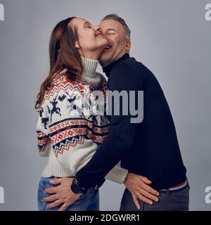 Homme adulte embrassant sa femme heureuse en riant. Couple aimant en chandails chauds posant pour un portrait de famille. Hiver vacances photoshoot concept en studio Banque D'Images