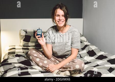 Thème diabète. Jeune femme caucasienne à la maison dans la chambre à coucher sur le lit utilise la technologie de l'appareil pour mesurer le sang gl Banque D'Images