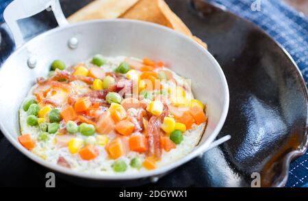 Petit-déjeuner avec des œufs frits dans une poêle avec du bacon et des légumes sur une assiette foncée. Banque D'Images