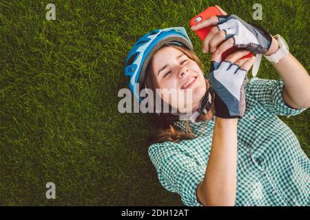 Thème sport et technologie. Belle femme caucasienne étudiant avec un sourire Toothy repose dos herbe verte de repos dans le casque de vélo hea Banque D'Images