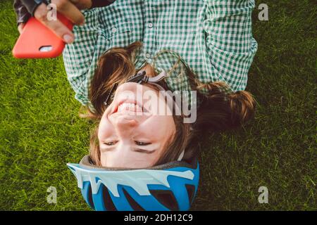 Thème sport et technologie. Belle femme caucasienne étudiant avec un sourire Toothy repose dos herbe verte de repos dans le casque de vélo hea Banque D'Images