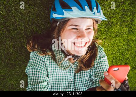 Thème sport et technologie. Belle femme caucasienne étudiant avec un sourire Toothy repose dos herbe verte de repos dans le casque de vélo hea Banque D'Images