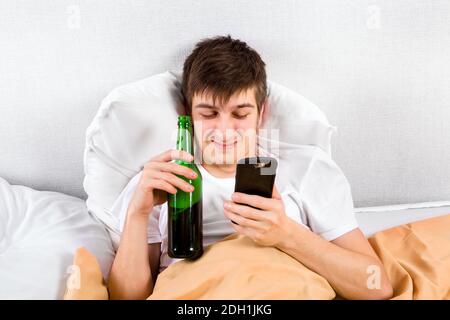 Un jeune homme mécontent avec un téléphone et une bouteille de bière Dans le lit Banque D'Images