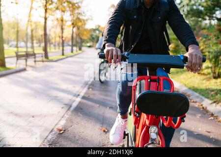 Gros plan photographique du corps d'un garçon étudiant avec un sac à dos sur son dos portant un vélo électrique rouge partagé un sentier de parc bordé d'arbres au coucher du soleil Banque D'Images