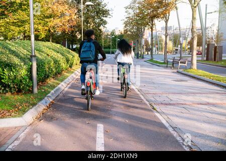 caméra arrière deux jeunes hommes et une femme à la conduite d'un piste cyclable avec un vélo électrique partagé dans un beau parc avec de nombreux arbres au coucher du soleil Banque D'Images