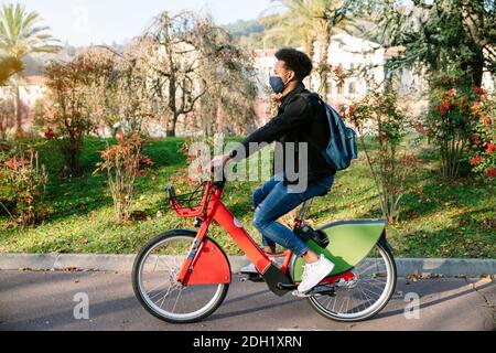 jeune homme un étudiant marocain à vélo électrique partagé dans un beau parc avec de nombreux arbres au coucher du soleil, portant un masque pour le pand du coronavirus Banque D'Images