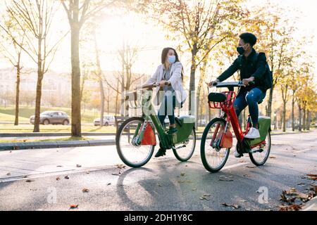 avant de deux jeunes homme et femme, le long d'un piste cyclable avec un vélo électrique de partage dans un beau parc avec de nombreux arbres au coucher du soleil portant un f Banque D'Images