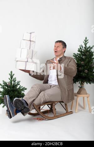 Portrait horizontal de Noël d'un homme de métier habillé élégant et décontracté, assis sur un traîneau jonglant, des boîtes cadeau enveloppées de blanc, le tout sur un fond blanc. Banque D'Images