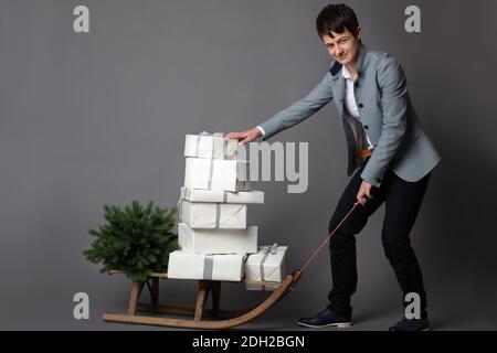 Portrait horizontal de Noël d'une femme cadre chic et décontractée tirant un traîneau rempli de boîtes-cadeaux blanches et argentées sur un fond gris. Banque D'Images