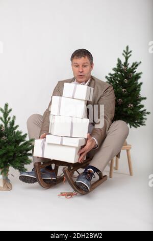 Portrait horizontal de Noël d'un homme de métier habillé élégant et décontracté, assis sur un traîneau jonglant, des boîtes cadeau enveloppées de blanc, le tout sur un fond blanc. Banque D'Images