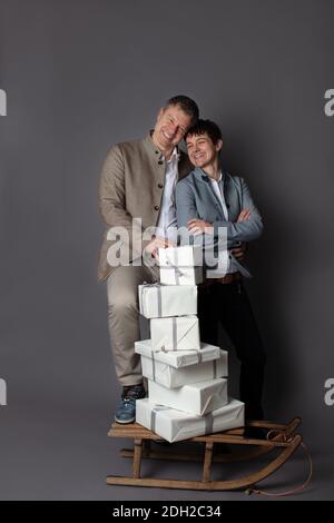 Thème de Noël vertical couple portrait habillé élégant décontracté dans bleu, blanc, beige pantalon et blazers sur fond gris. Au premier plan un traîneau en bois avec une pile de boîtes-cadeaux blanches. Banque D'Images