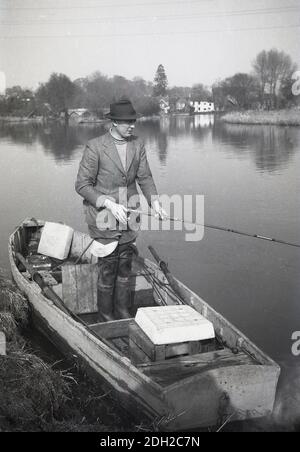 Années 1950, historique, sur une rivière anglaise calme, une photo d'un gentleman pêchant. Debout dans un petit bateau en bois au bord de la rivière, et portant de longues bottes wellington ou des échassières, une veste à carreaux et un chapeau, avec canne à pêche à la main, Angleterre, Royaume-Uni. Banque D'Images