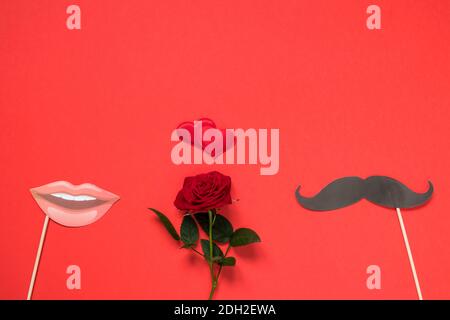 Saint-Valentin, amour, concept romantique. Fleur de rose rouge fraîche et coeur rouge avec moustaches et lèvres prop papier sur fond rouge. Banque D'Images