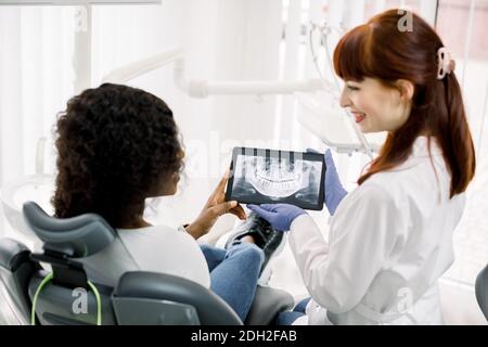 Jeune femme africaine assise en chaise de dentisterie et regardant la radiographie panoramique. Jolie femme médecin explique les manières de traitement ou Banque D'Images