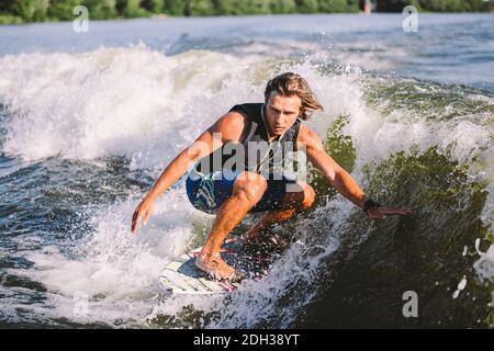 Belle wakesurf blonde aux cheveux longs en gilet à bord le long des vagues du lac. Un athlète sportif se réveille en surfant sur la rive en été Banque D'Images