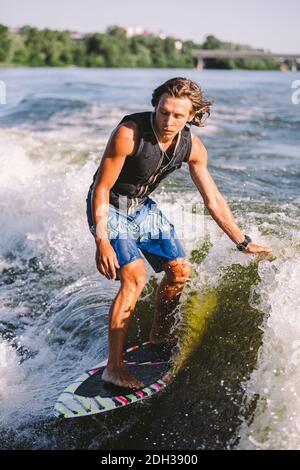 Belle wakesurf blonde aux cheveux longs en gilet à bord le long des vagues du lac. Un athlète sportif se réveille en surfant sur la rive en été Banque D'Images