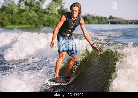 Belle wakesurf blonde aux cheveux longs en gilet à bord le long des vagues du lac. Un athlète sportif se réveille en surfant sur la rive en été Banque D'Images