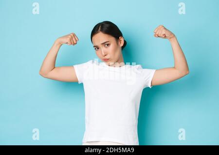 Sassy jolie fille asiatique en t-shirt blanc, montrant sa force, l'entraînement en salle de gym, Flex biceps et regardant confiant, l'encourager Banque D'Images