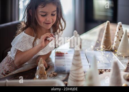 Jolie petite fille avec cheveux bouclés faire des métiers et décorer le cône de sapin de Noël avec des boutons. Banque D'Images