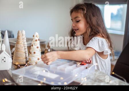 Jolie petite fille avec cheveux bouclés faire des métiers et décorer le cône de sapin de Noël avec des boutons. Banque D'Images