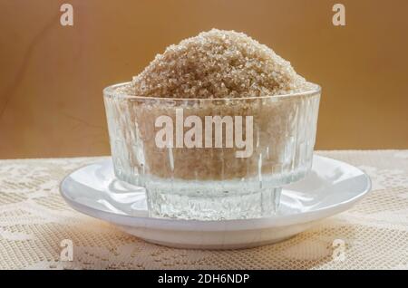 Un petit plat transparent qui se trouve dans une petite assiette blanche sur une table recouverte de nappe lacey est rempli de cristals de sucre de canne. Banque D'Images