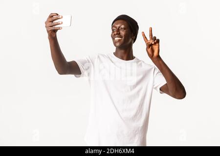Portrait de beau et joyeux Black Guy dans le bonnet, prenant selfie sur le téléphone mobile et montrant le geste de paix heureux, souriant plus de W Banque D'Images