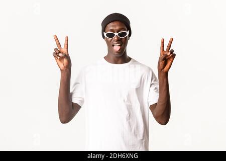 Portrait du jeune homme afro-américain drôle et optimiste en bonnet et lunettes de soleil, montrant des gestes de paix et la langue adhérante, wh Banque D'Images