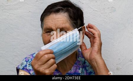 Une femme âgée froissée met un masque médical de protection en touchant son oreille avec les doigts. La vieille dame montre comment porter le masque de manière appropriée. Peopl Banque D'Images