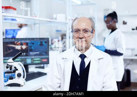 Scientifique senior assis sur son lieu de travail pendant des expériences médicales. Scientifique âgé portant un blouse de laboratoire travaillant pour développer un nouveau médical en vacances avec un assistant africain en arrière-plan. Banque D'Images