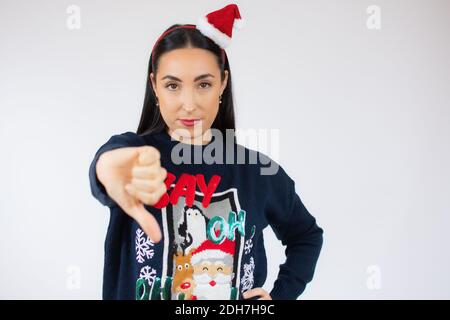 Jeune belle femme brune portant chapeau de père noël debout sur blanc arrière-plan à l'air malheureux et en colère montrant le rejet et négatif avec le pouce Banque D'Images