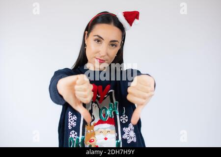 Jeune belle femme brune portant chapeau de père noël debout sur blanc arrière-plan à l'air malheureux et en colère montrant le rejet et négatif avec pouces d Banque D'Images