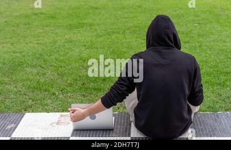 Un jeune homme portant un sweat à capuche assis dans un parc avec son ordinateur portable Banque D'Images