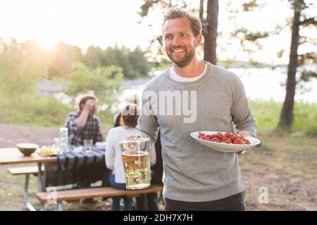 Joyeux homme tenant une carafe à bière et des fraises avec des amis assis à la table de pique-nique en arrière-plan Banque D'Images