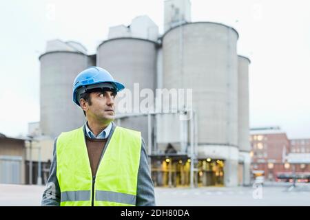 Homme ouvrier regardant loin en se tenant contre des silos de ciment Banque D'Images