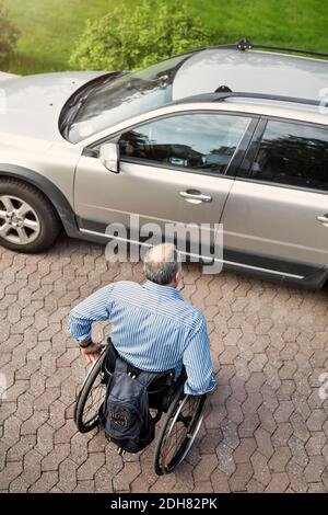 Vue en grand angle de l'homme adulte en fauteuil roulant se déplaçant vers voiture dans la rue Banque D'Images