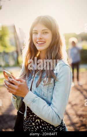 Portrait d'une adolescente heureuse tenant un smartphone debout au parc Banque D'Images